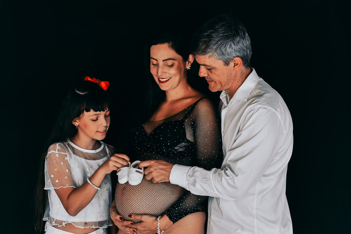 Pai, mãe e filha segurando sapatinhos de bebê na barriga